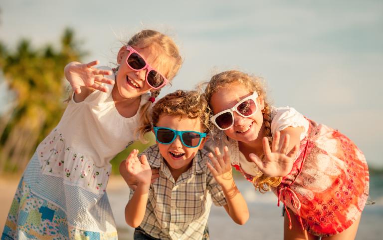 Three Children on a Beach all Wearing Sunglasses Smiling and Waving