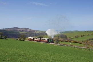 A steam train going along the track in between green fields of the North Devon coastline on a bright sunny day